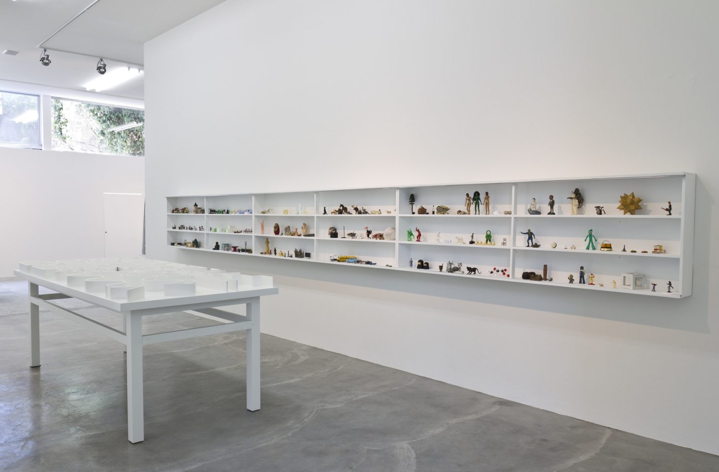 An installation photo of Pedro Reyes' Museum of Hypothetical Lifetimes. A table and shelves are installed in a bright, white gallery. Lots of small objects are arranged on the shelves.