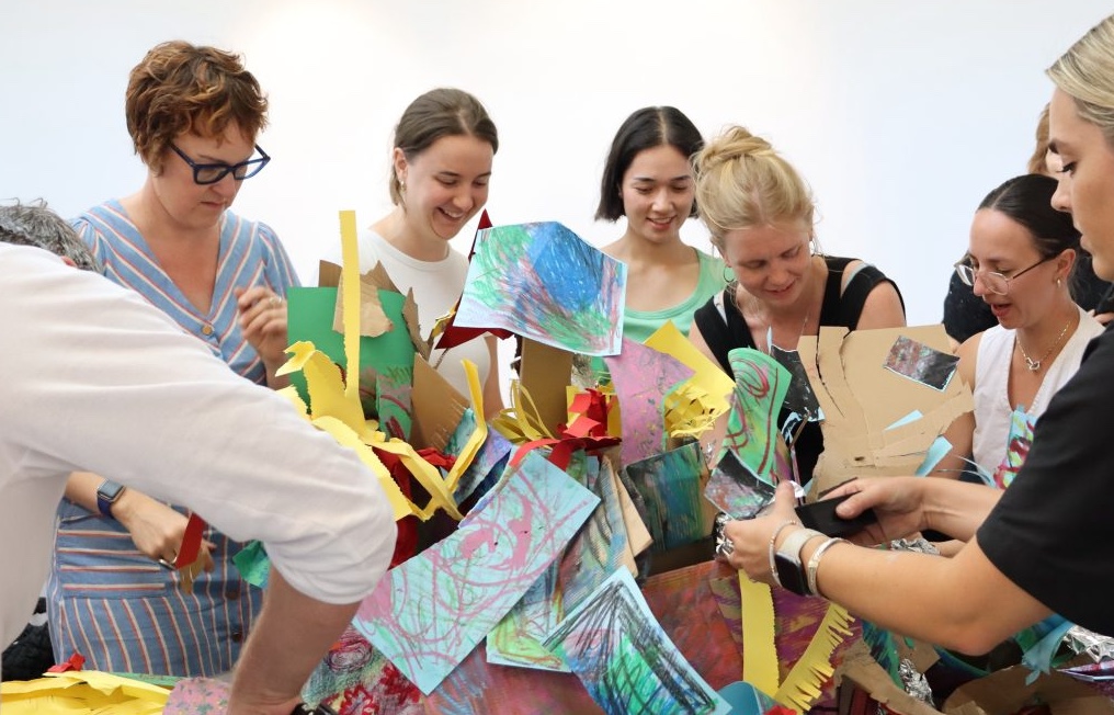 A group of teachers sit around tables in a bright gallery space at South London Gallery, taking part in a creative professional development session led by artist Yolanda Harris in 2025. Art materials, notebooks and drinks are on the tables, and participants are talking and smiling as they work together.