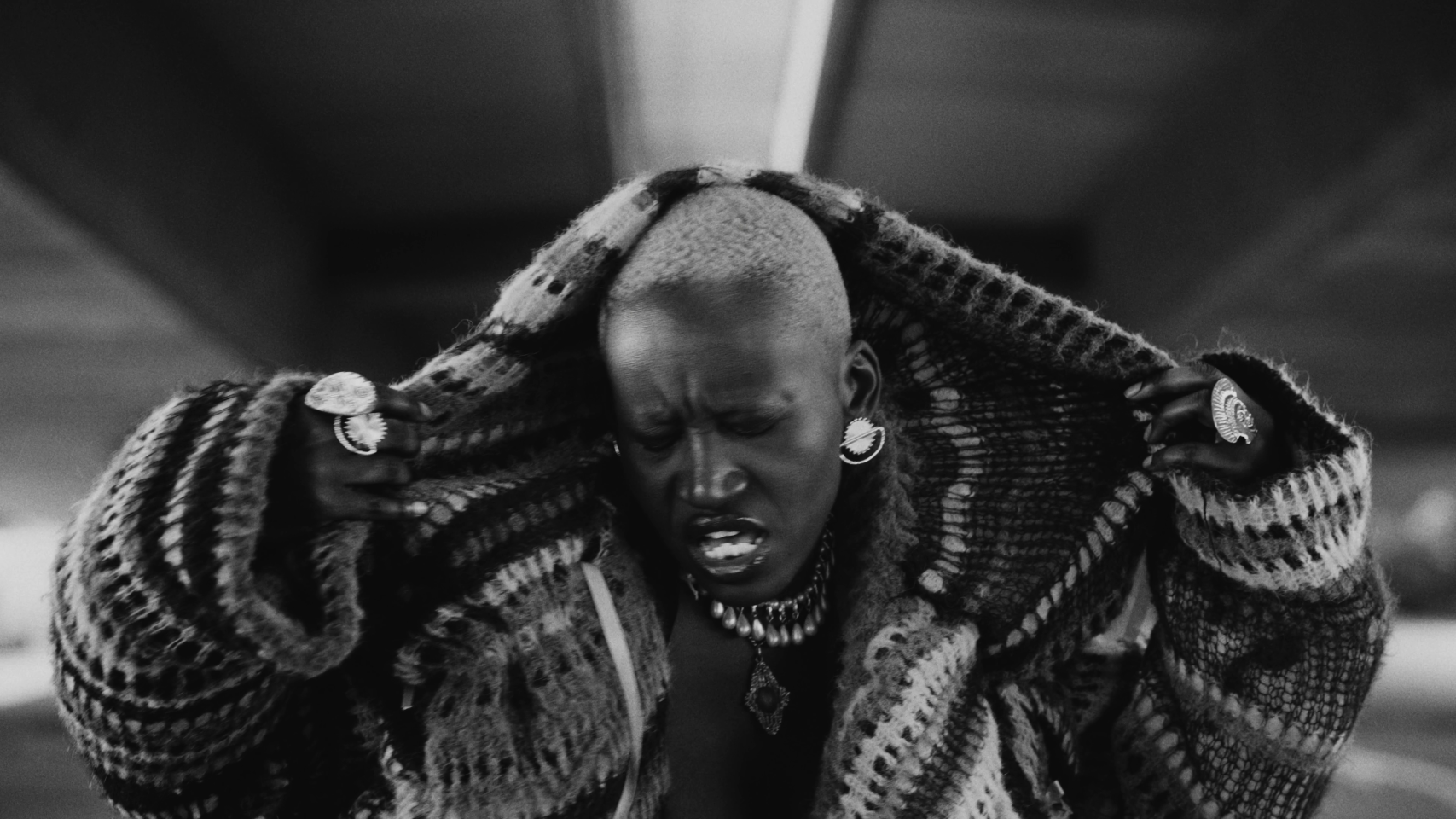 A black and white still from a music video. A woman with a shaved head wearing a knitted hoody pulls it over her head.
