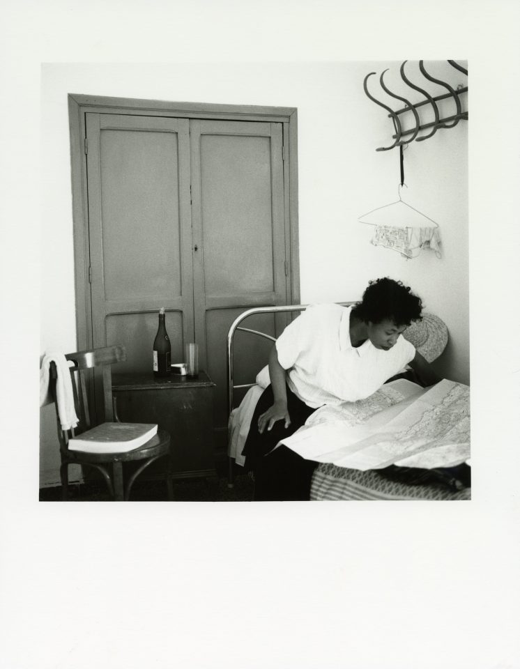 A photography self portrait by artist Joy Gregory. The black and white image shows the artist, wearing white, in a hotel room.