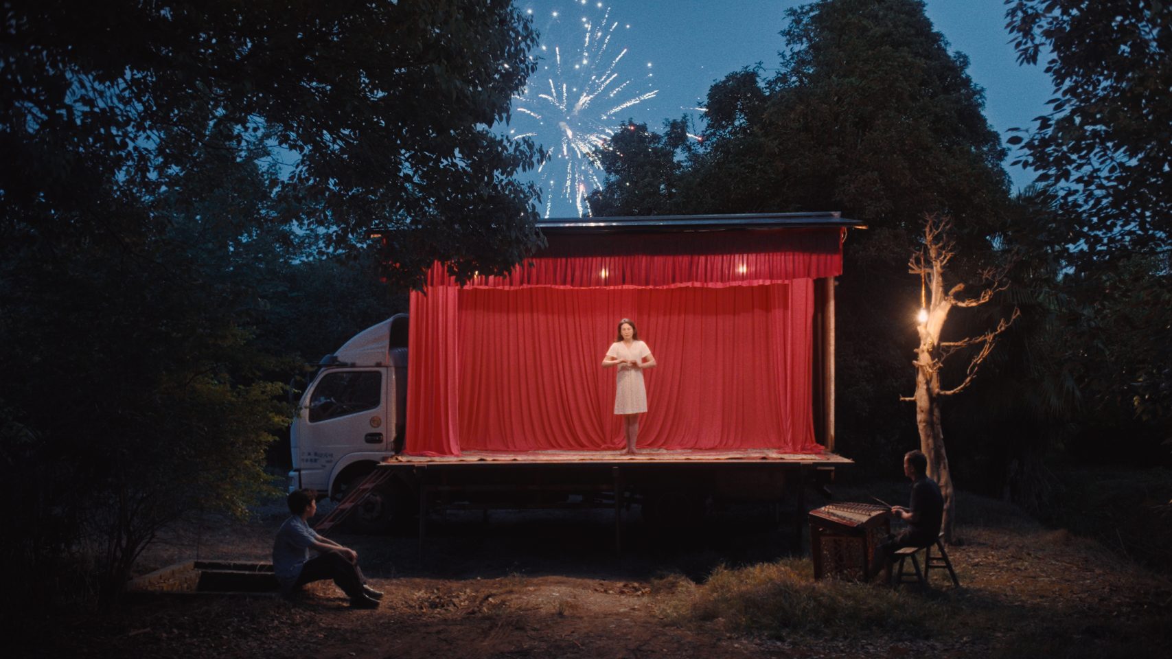 A figure stands on a red stage. The stage is set in the back of a truck parked in a wood.