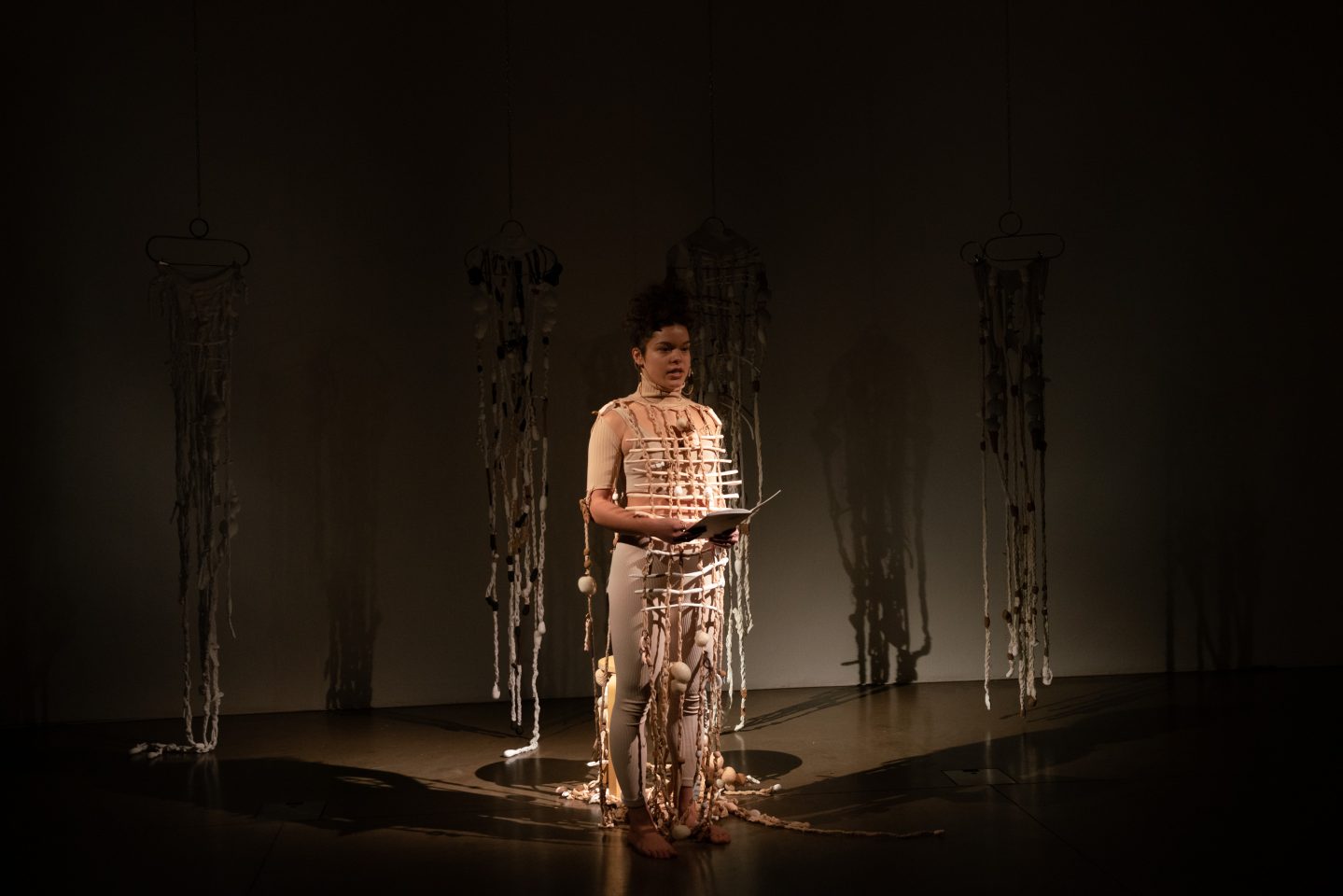 An artist does a performance piece in a dark room. They are spotlit, and wear a beige costume and hold a book in front of them.