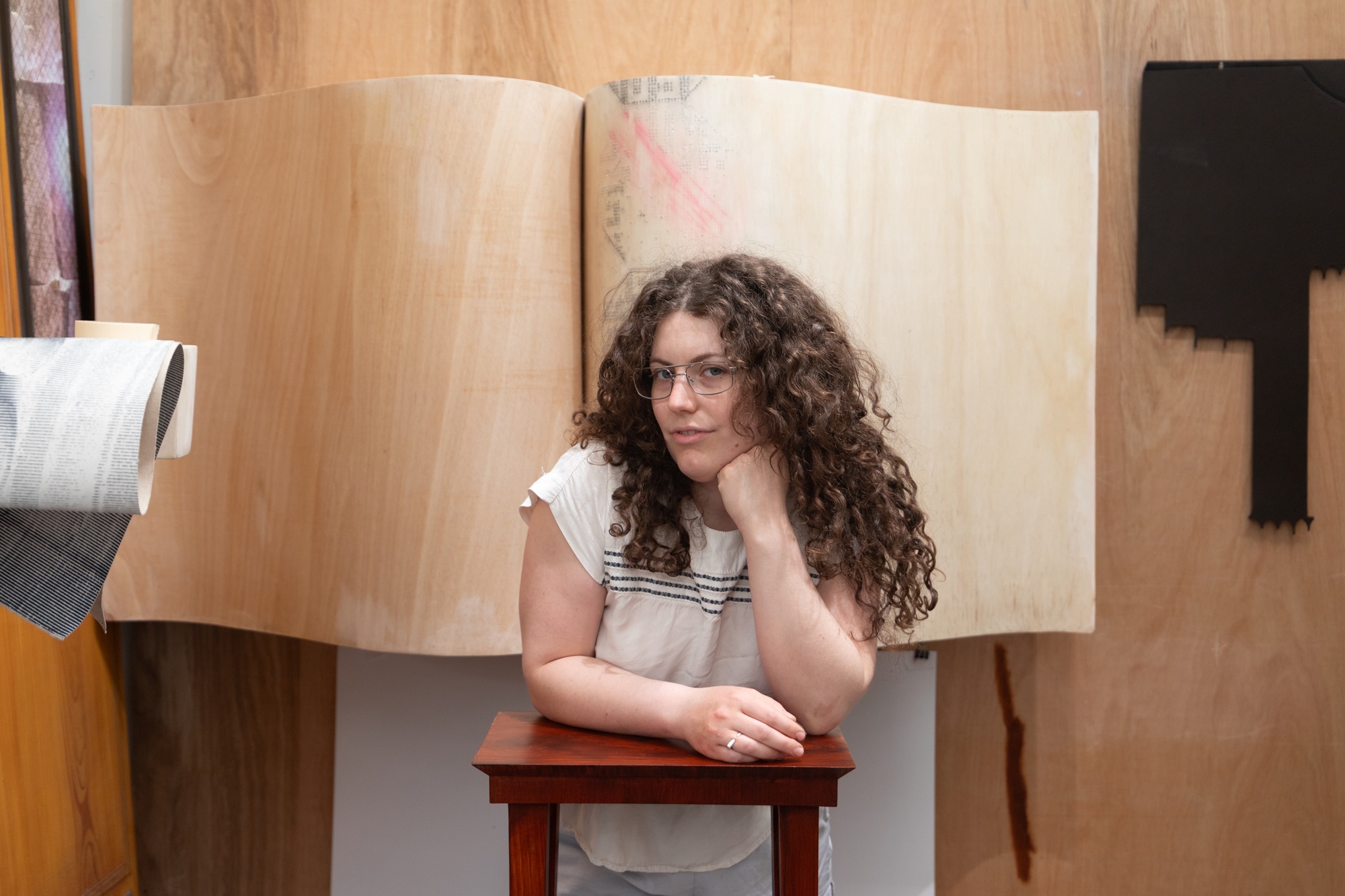 A photo of artist Maya Silverberg in her studio. She leans on a wooden table in front of wooden sculptures.