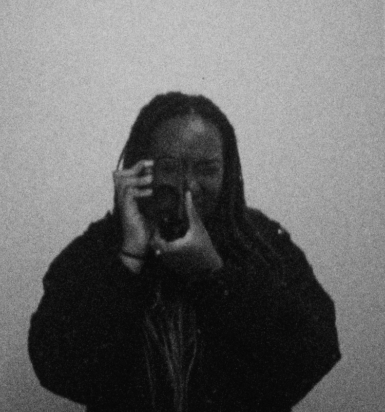 A photo of artist Ufuoma Essi holding a camera up to her face as if she is taken a photo.