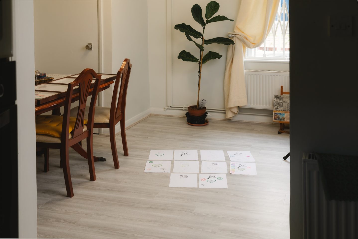 A living room with pale wooden flooring, viewed through a doorway. On the floor are sheets of paper with simple drawings and laid out in a grid. To the left is a wooden dining table with chairs, and to the back is a potted indoor plant near a window with light curtains.