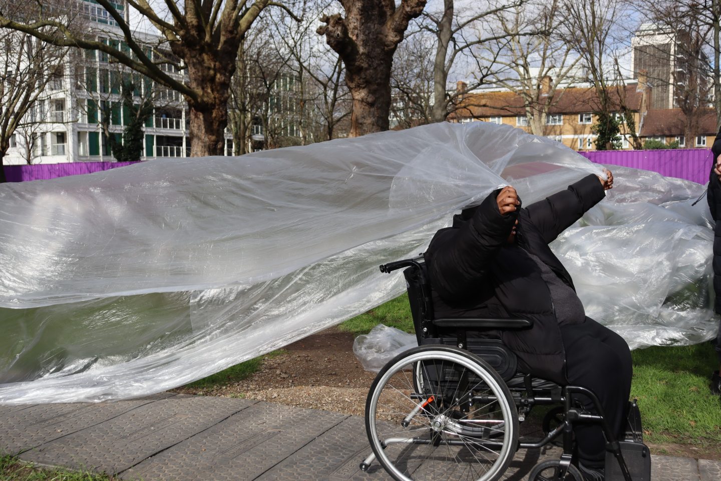A person holds up plastic tarp outside. There are buildings in the background.