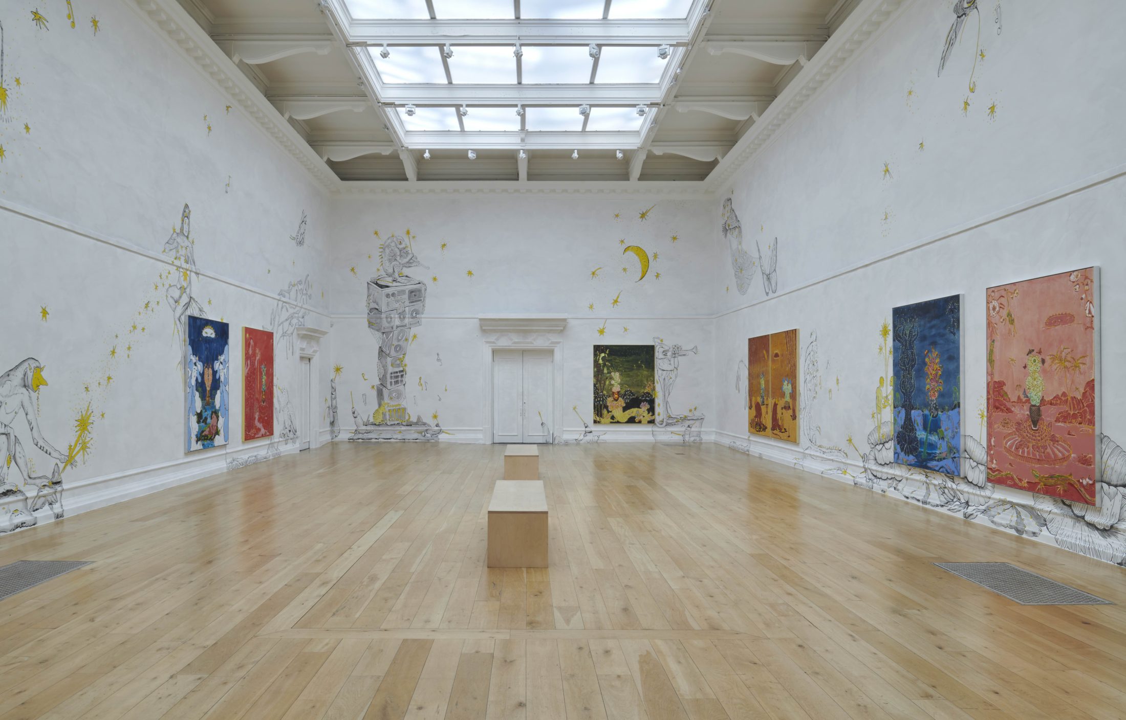 Wide view of a bright gallery space with a skylight ceiling and wooden floors. The white walls are covered with whimsical, hand-drawn mural elements in black and yellow—featuring fantastical creatures, musical motifs and star-like bursts. Large, colourful paintings hang along both sides of the room. A simple wooden bench sits in the centre, and double doors are visible at the far end.