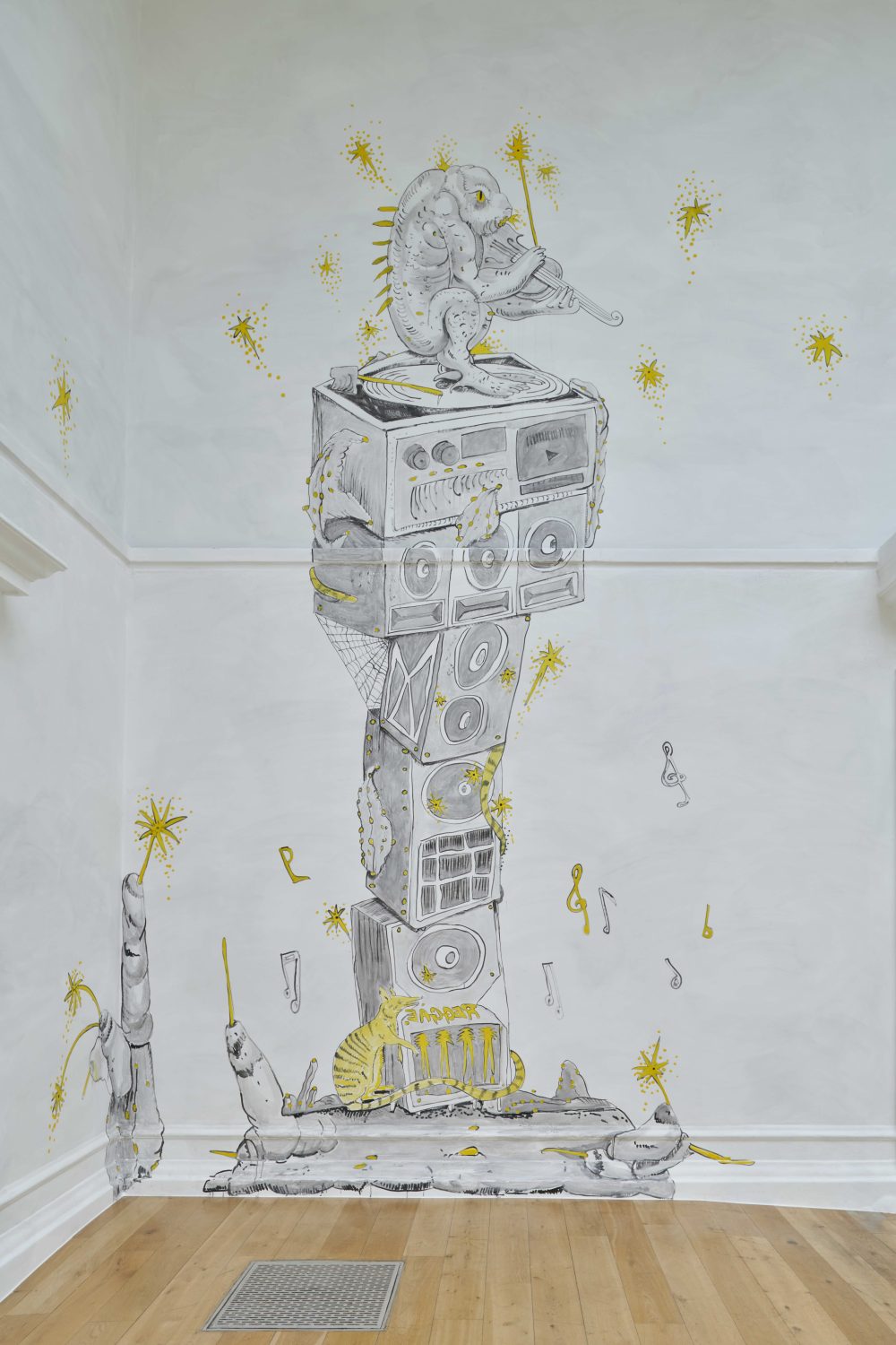 A large wall drawing in a gallery space shows a whimsical, stacked totem of objects and creatures. At the top, a small cartoon-like creature plays a stringed instrument while standing on a turntable. Below, stacked speakers and abstract forms create a vertical column, with a cat-like figure near the base. Yellow accents highlight stars, sparks and details across the monochrome illustration. The mural sits on a pale grey wall above a wooden floor.