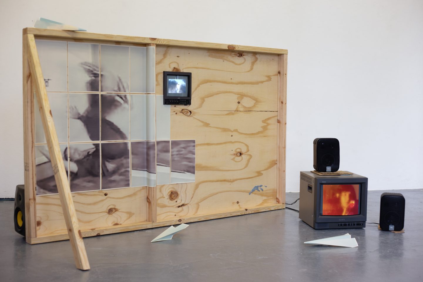 Mixed-media installation in a gallery space featuring a wooden, box-like panel propped open on the left, with a grid of semi-translucent paper images showing a blurred figure. A small monitor is embedded in the wood displaying cloudy sky footage, while to the right a CRT television on the floor shows a close-up of a face in warm tones, flanked by two small speakers. Sheets of folded paper lie scattered on the concrete floor.