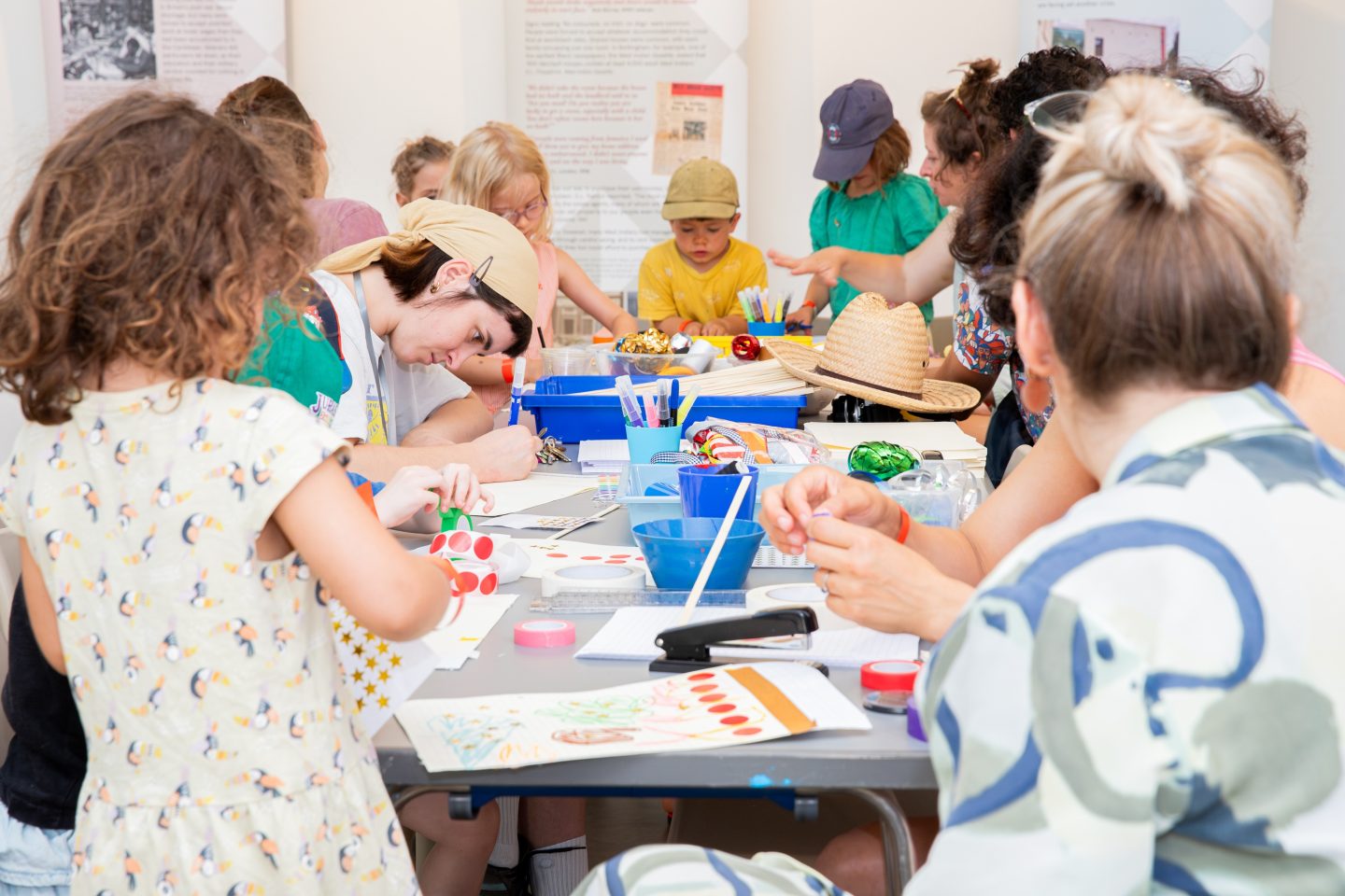 A large group of children and adults gathered around a long table participating in a hands-on arts and crafts activity. Participants are drawing, coloring, and decorating paper projects using markers, stickers, tape, and other craft supplies spread across the table. Blue containers hold art materials, and completed and in-progress drawings are visible in the foreground. The activity takes place indoors in a bright room with informational display panels on the walls in the background