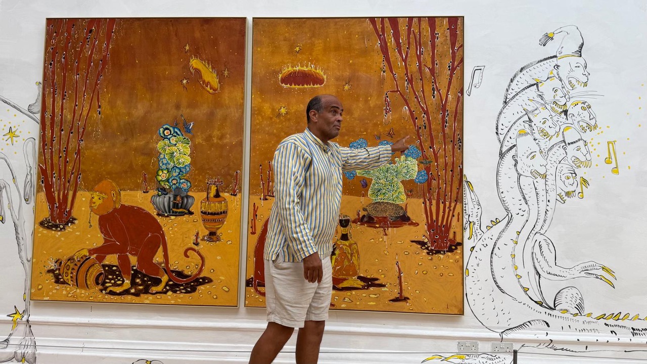 Man stands in front of two large yellow paintings, pointing and signing at the pieces of work.