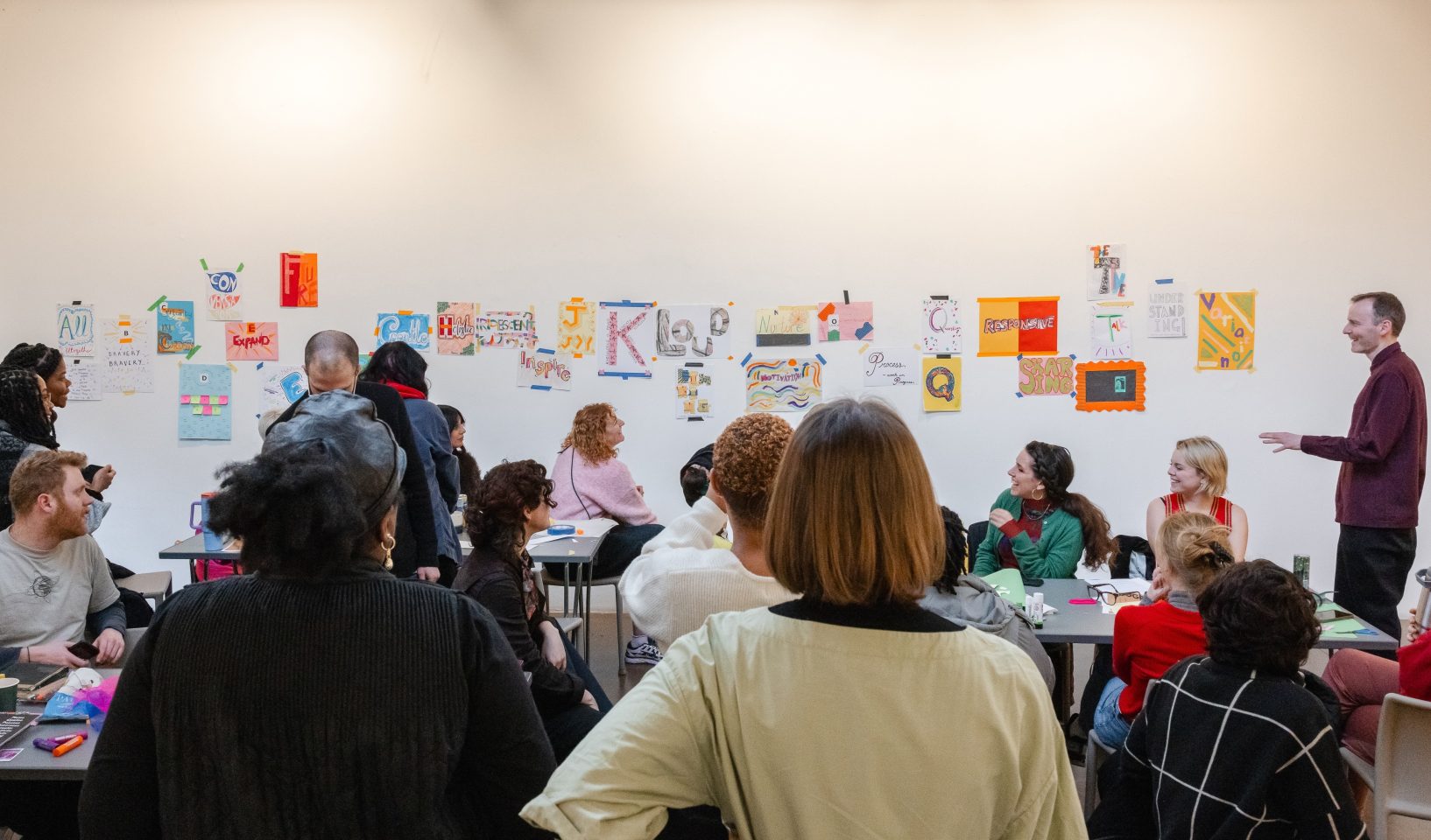 People gathered in a workshop space, seated and standing around tables, while a facilitator speaks beside a wall displaying colourful handmade posters and creative prompts.