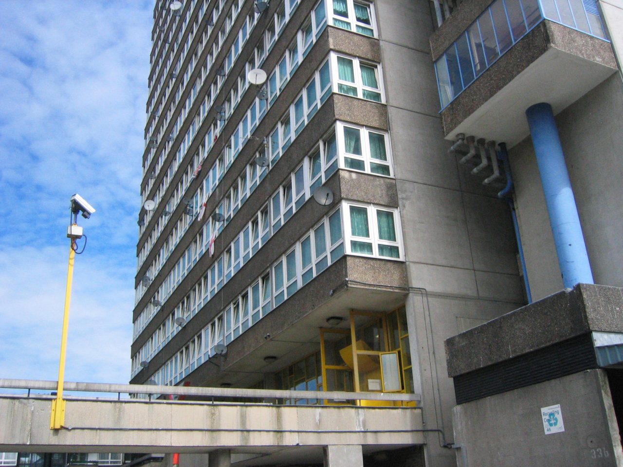 Tall residential tower block with rows of windows and satellite dishes, viewed from below with a CCTV camera in the foreground against a blue sky.