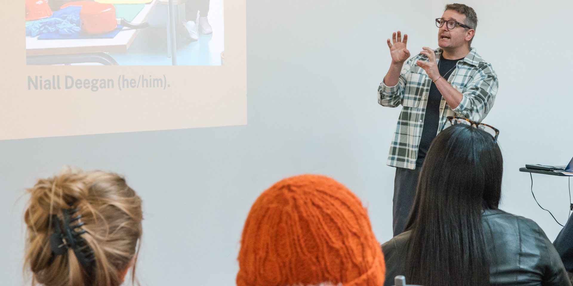 A speaker stands at the front of a presentation room, gesturing while addressing an audience seated in the foreground. A projected slide is visible on the wall beside the speaker, showing a photograph and text. Several attendees are seen from behind, facing the presentation in a bright, minimalist gallery setting.