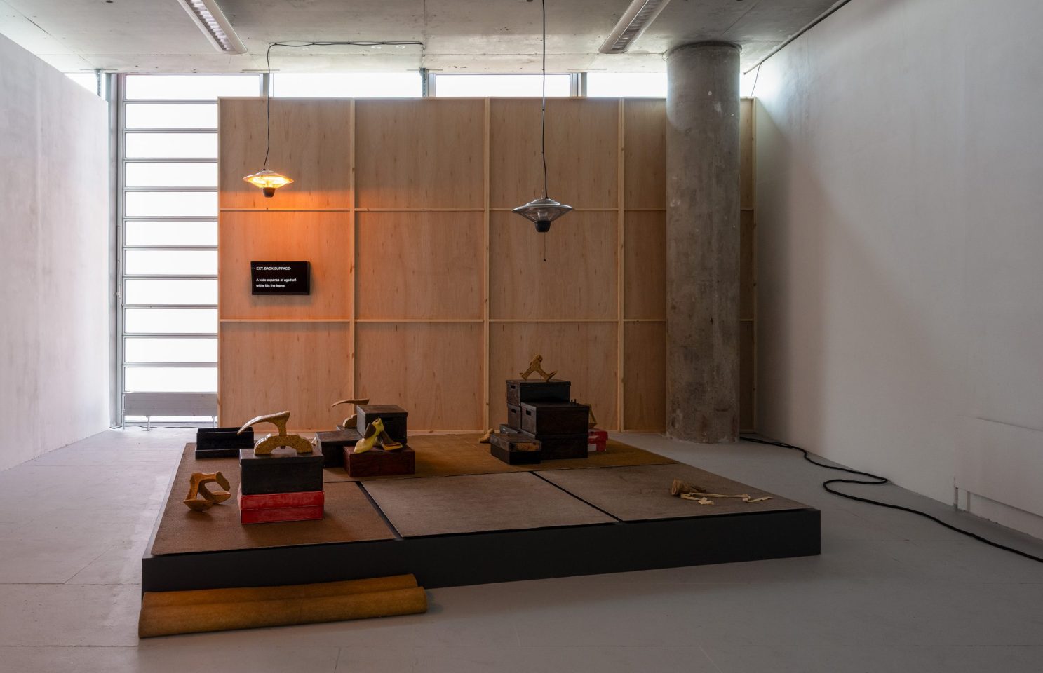 A piece of installation art consisting of a temporary wooden wall and an assortment of objects on a wooden plinth.